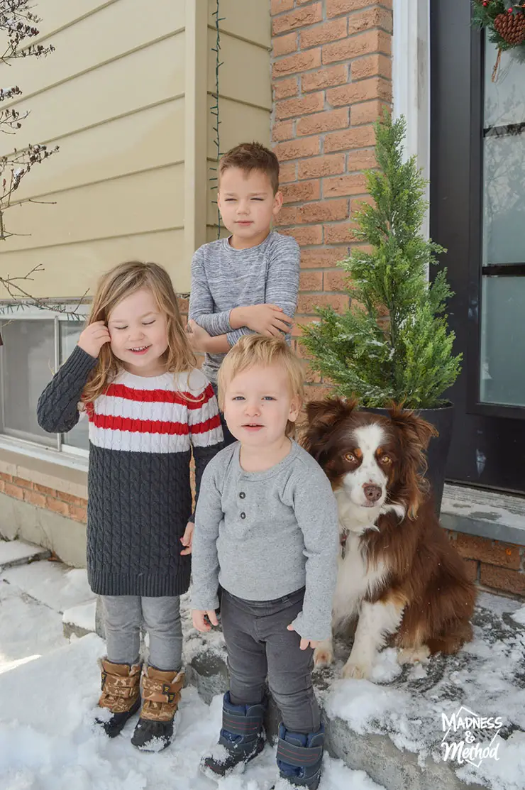 three young kids and dog standing on snow covered porch with small faux evergreen tree